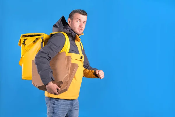 Unshaven caucasian young blond guy food delivery guy in branded yellow uniform is walking and holding boxes of pizza from restaurant, late with delivery to customer filmed on blue background.