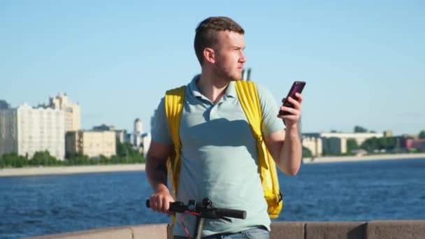 Food delivery man courier with yellow thermos bag and electric scooter, lost in city and uses navigation in phone looking for address of food delivery from restaurant, talking on phone with customer — Stock Video