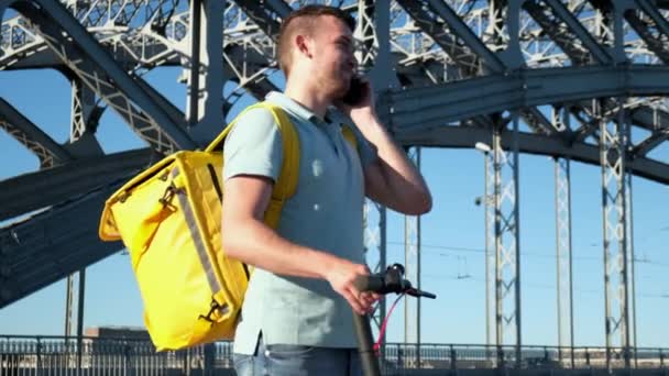 Courier entrega de alimentos com térmica amarela e uma scooter elétrica, na rua contra o fundo da ponte falando por telefone sorrindo e à procura de endereço de entrega de alimentos a partir de restaurante. — Vídeo de Stock