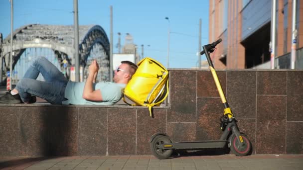 Unrasierter blonder kaukasischer Mann mit Sonnenbrille Essenslieferant mit gelber Thermoskanne und Elektroroller, liegend und liegend beim Spielen auf dem Handy auf der Straße vor dem Hintergrund der Brücke — Stockvideo