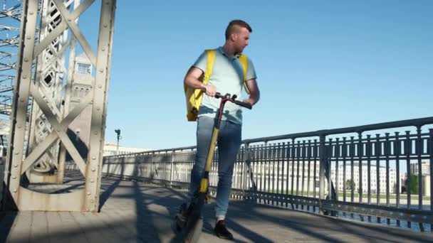 Jonge blanke man die eten aflevert van restaurant rijdt op een elektrische scooter over de brug in de stad en praat over de telefoon, op zoek naar geolocatie coördinaten van de plaats van levering. koerier voor snelle levering — Stockvideo