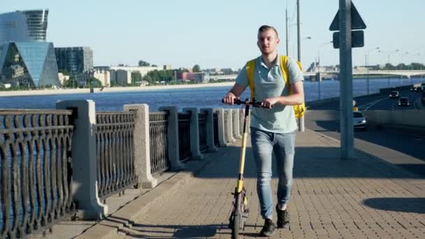 Jovem entregando alimentos e produtos de restaurantes e supermercados com saco de geladeira amarelo em suas costas caminha pela cidade e segura uma scooter elétrica em suas mãos e olha em volta . — Vídeo de Stock