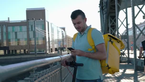 Comida de mensajería de restaurantes y supermercados con bolsa de termo y una scooter eléctrica mira el teléfono y busca la dirección de entrega y habla por teléfono en el puente de hierro en la ciudad durante el día — Vídeos de Stock