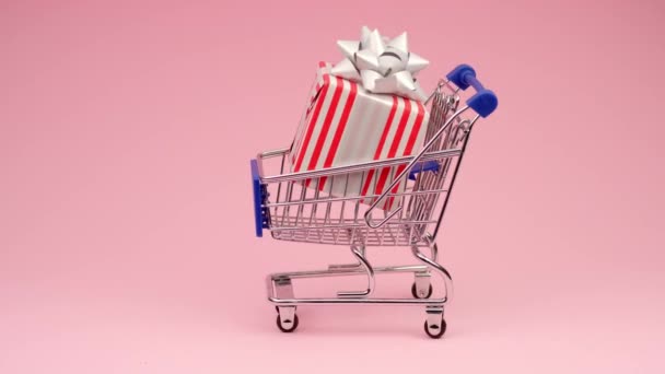 Concepto de compra de bienes en tiendas online y productos en supermercados. Mano de hombre empuja pequeño carro de supermercado con caja de regalo completa sobre fondo rosa con espacio para el texto. VENTA grande en tienda . — Vídeo de stock