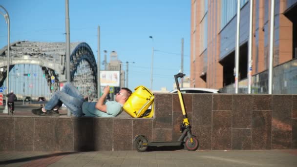 El repartidor de comida de los restaurantes de la ciudad, el mensajero miente y descansa en el banco de la ciudad jugando juegos por teléfono, el mensajero recibe una orden por teléfono y rápidamente sale en una scooter eléctrica al restaurante — Vídeos de Stock