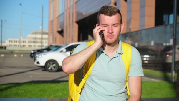 Homem alegre e sorrindo entrega de alimentos com saco senta-se perto de construção de negócios e fala ao telefone com o comprador de alimentos, olhando ao redor com a cabeça para o endereço certo. Entrega rápida na cidade de restaurante . — Vídeo de Stock