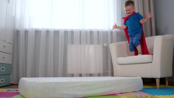 Små barn i rött Super man kid super hjälte Cape hoppar från stol till madrass ligger i barnrum. Fall av superhjälte, pojkar misslyckades försök att flyga, pojkar fantasi. Långsam rörelse video — Stockvideo