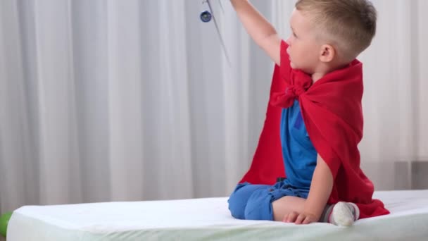 Niño rubio caucásico super héroe niño está jugando con el avión de juguete en la habitación de los niños vestidos con impermeable rojo y camiseta azul. Niño sueña con convertirse en piloto aéreo de un avión. — Vídeo de stock