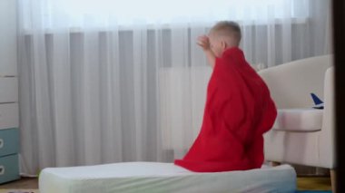 Ağır çekimde sarışın, beyaz kahraman çocuk. Kırmızı kahraman paltosu içinde. Eğleniyor, trambolinde olduğu gibi yatakta zıplıyor.