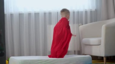 Ağır çekimde sarışın, beyaz kahraman çocuk. Kırmızı kahraman paltosu içinde. Eğleniyor, trambolinde olduğu gibi yatakta zıplıyor.