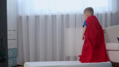 Ağır çekimde sarışın, beyaz kahraman çocuk. Kırmızı kahraman paltosu içinde. Eğleniyor, trambolinde olduğu gibi yatakta zıplıyor.