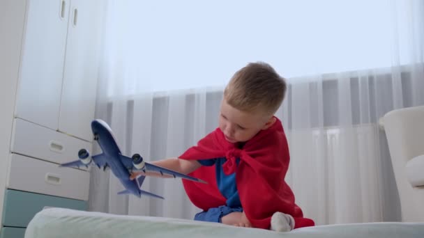 Blond kaukaski chłopiec super bohater dzieciak bawi się samolotem zabawki w pokoju dla dzieci ubrany w czerwony płaszcz przeciwdeszczowy i niebieski t-shirt. Chłopiec marzy o zostaniu pilotem samolotu.. — Wideo stockowe