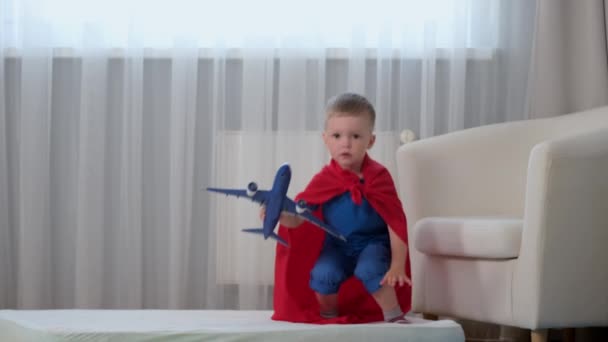 Powolny ruch blondynka biały chłopiec super bohater dziecko zabawy z zabawka samolot, skakanie na materacu, trampolina z zabawką w ręku ubrany w czerwony płaszcz przeciwdeszczowy bohatera i niebieski t-shirt w pokoju dla dzieci. — Wideo stockowe