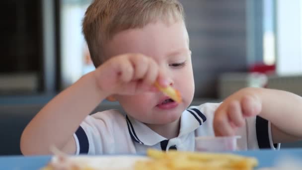 Portret małego blondyna białego chłopca w białej koszulce siedzi przy stole w restauracji fast food i je niezdrowe jedzenie. Dziecko zanurza frytki w ketchupie, wkłada do ust i żuje jedzenie.. — Wideo stockowe