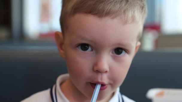 Cute blond caucasian boy portrait drinks sweet soda through tube and puts white paper cup on table in fastfood restaurant. Quick snack in fast food, quench your thirst with cold soda. — Stock Video