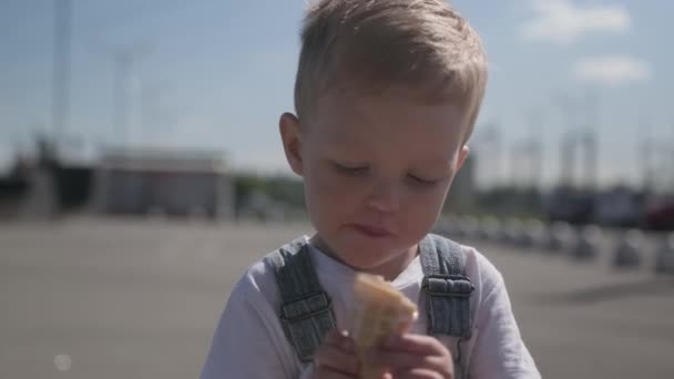 Retrato de chico rubio caucásico en camiseta blanca y overoles de mezclilla, comiendo y disfrutando de helado cremoso frío en taza de cono de gofre, sentado afuera en clima soleado. — Vídeo de stock