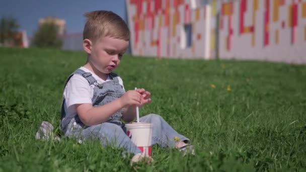 St.Petersburg,ロシア, 2020年8月:外の夏の天気で青い草の上に座って、わらパイプと白い紙カップから冷たい飲み物を飲むことによって彼の渇きを癒すブロンドの白人少年. — ストック動画