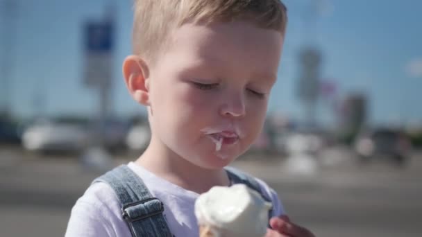 Portret białego chłopca w białym t-shircie i dżinsowych kombinezonach, jedzącego i cieszącego się kremowymi zimnymi lodami w waflowym kubku, siedzącego na zewnątrz w słoneczną pogodę. — Wideo stockowe