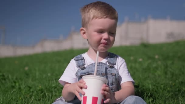 Blond caucasian boy sits on green grass in summer weather outside and quenches his thirst by drinking cold drink from straw pipe and white paper Cup. Child drinks coke with ice in summer heat of sun. — Stock Video