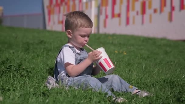 Cute blond boy sits on green grass and quenches his thirst in summer Sunny hot weather. Child drinks cold and refreshing soda cola from white paper cup through tube in hot heat city. — Stock Video