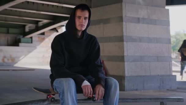Portrait young serious not smiling caucasian skateboarder, looking at camera. Guy is sitting on skateboard in black hoodie under an iron bridge on skate Park. Slow motion orbit shot. — Stock Video