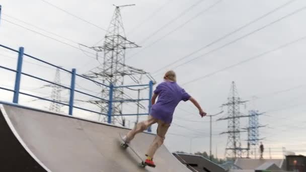 Teenager mit langen blonden Haaren fährt Skateboard und macht verschiedene Tricks. Skateboarderfahrten auf der Rampe machen bei trübem Wetter Tricks auf dem Skateboard im Skatepark. Ein extrem traumatischer Sport — Stockvideo