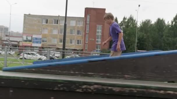 Mladý blonďatý chlapec skateboardista jede dopředu na skateboardu v skate parku v oblačném počasí. Profesionální bruslař teen zabývající se extrémními sporty v skatepark představení triky. — Stock video