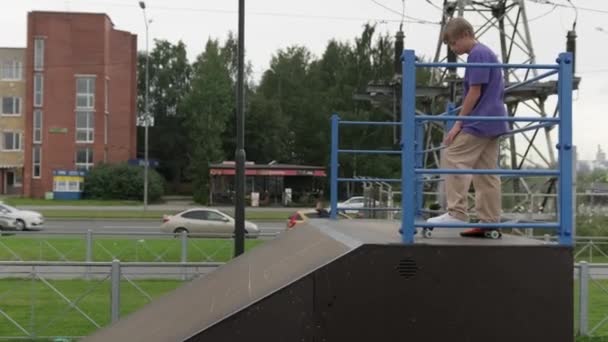 Fiatal szőke fiú gördeszkás lovagol előre gördeszkán a gördeszka parkban felhős időben. Profi korcsolyázó tini részt extrém sportok skatepark bemutató trükkök. — Stock videók