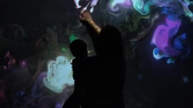 Çocuklu bir kız interaktif video kurulumuyla oynuyor. Yeni sanat formu, üretken grafikler. Çocuklu kız silueti çok renkli boyalar çiziyor etkileşimli kurulum.