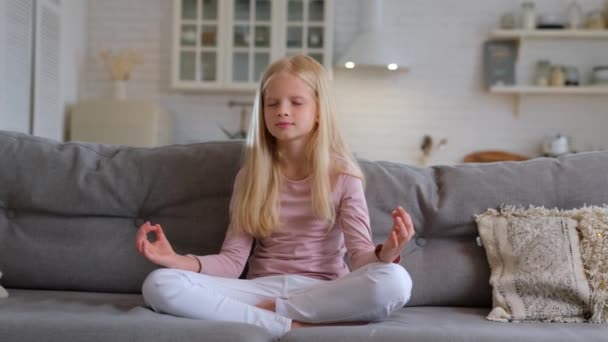 Ragazza caucasica bionda fa yoga al mattino, si siede sul divano in appartamento in posizione loto e medita con gli occhi chiusi durante il giorno. Attraente ragazza snella rilassa immerge nel mondo scoperta di sé — Video Stock