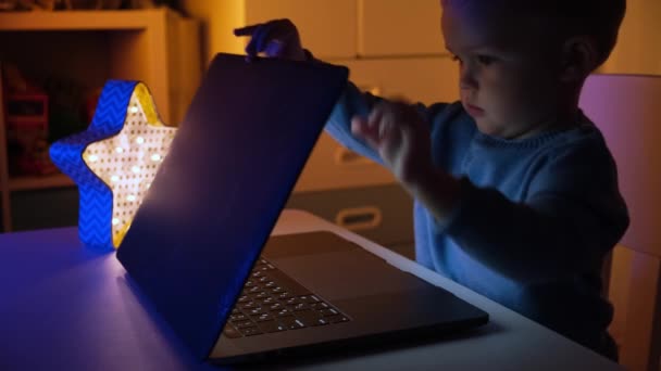 Ребенок открывает ноутбук и прижимает руки к клавиатуре в темной комнате, играет в игры на ноутбуке, мальчик просматривает интернет-пространства для ноутбука. Крупный план сосредоточенный мальчик пытается выполнить трудную задачу во время учебы — стоковое видео