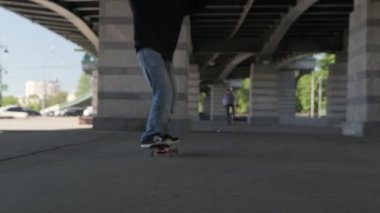 Gençler kaykayın ön tekerleklerinde kaykay parkında demir bir köprünün altında denge numaraları yapıyorlar. Koyu renk elbiseli profesyonel kaykaycı kaykayla numara yapıyor.