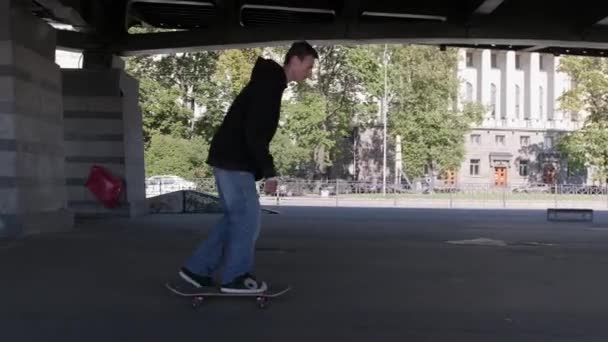 Tiener rijdt op voorwielen van skateboard, het uitvoeren van balancing trucs op de site van skate park onder een ijzeren brug. Professionele skateboarder kerel in donkere kleding voert truc op skateboard — Stockvideo