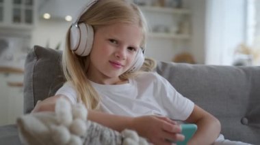 Kafasında kulaklıklarla genç sarışın güzel bir kızın portresi ve elinde telefonla oturma odasındaki kanepede oturup müzik dinleyerek ve eğlenerek. Kız kulaklıkla şarkı dinliyor..