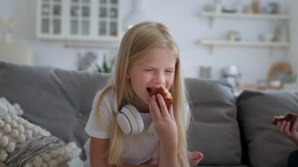 Портрет молодої блондинки красива дівчина з білими навушниками втамовує голод, тримає смачний свіжий кекс і їсть його, дивлячись на оператора камери, що сидить на дивані у вітальні . — стокове відео