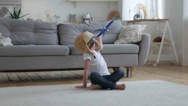 Счастливый маленький мальчик играет с белым пассажирским игрушечным самолетом в очках и кепке, мечтает летать над облаками, сидя днем на полу квартиры. Концепция путешествий и приключений. — стоковое видео