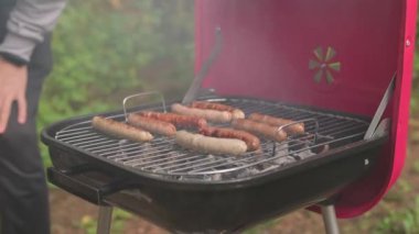 Piknikteki adam barbeküde sulu sosisler hazırlıyor. Aşçı, ızgara ızgaranın kapağını tavuk, domuz ve sığır sosisi ile açar ve ters çevirir. Kabuklu kızarmış sosisler parkta pişirilmiş kömürlerdir..