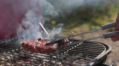 Ateş yanıyor, biftekli sığır filetosu pirzola barbeküde pişiyor. Şef tong kullanıyor ve eti ızgaraya çeviriyor. Orta boy ızgara, lezzetli biftek yakın çekim..
