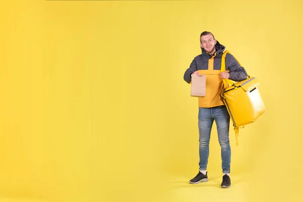 Cute caucasian food delivery courier smiles and looks ahead, holding food order in his hands and yellow refrigerator bag on yellow background hangs on his shoulder. Food delivery to home or office.