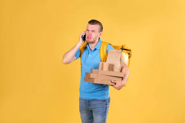 Delivery guy in blue uniform is walking talking on phone holding an order with food from restaurant and talking to customer behind his back he has delivery guy\'s refrigerator bag on yellow background.