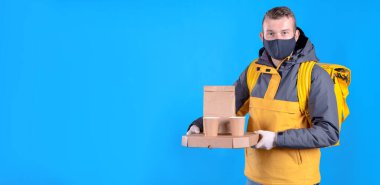 Siyah tıbbi maskeli ve lastik eldivenli genç kurye evinize ya da ofisinize yiyecek getiren ekolojik biyolojik çözünürlükte mavi arka planda paketlenmiş bir pizza siparişi tutuyor..