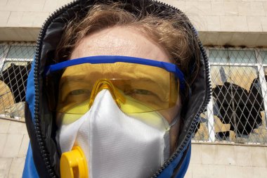 Koruyucu maskeli ve gözlüklü bir kadın ve evin arka planında bir kaput, koronavirüs konsepti, çevre felaketi konsepti