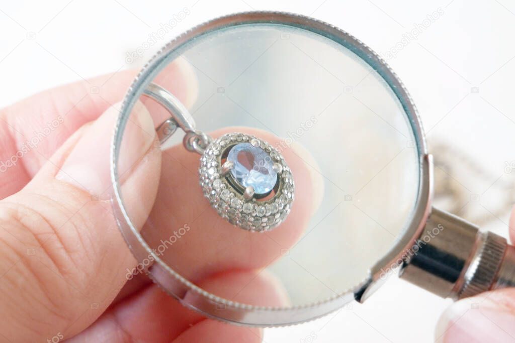 hand takes earring with big blue topaz and white diamonds around, jeweler looking at jewelry through magnifying glass, jewerly inspect and verify, pawnshop concept