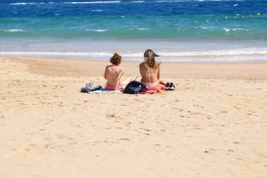 İki harika bronzlaşmış seksi bikinili çekici kız okyanusun yanındaki güneşli sropik plajda oturuyor.