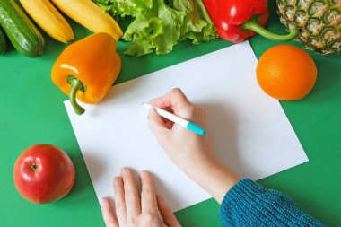 Taze organik sebzeler, meyveler ve beyaz boş kağıt tabaka. Biber, ananas, salatalık, elma, muz, yeşil arka planda boş defter ve kalem. Sağlıklı yiyecekler, beslenme planı ve sağlıklı yaşam konsepti. Üst görünüm