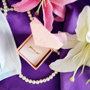 Düğün ve koronavirüs kavramı: mücevher kutusu, mavi sağlık maskesi, altın alyans ve pembe çiçek demeti.