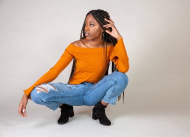 Studio bir beyaz arka plan çömelme poz vintage turuncu üst ve bol kot ile genç bir siyah kadın vurdu