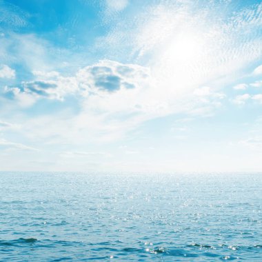 Mavi gökyüzünde güneş, denizin üzerinde bulutlar.