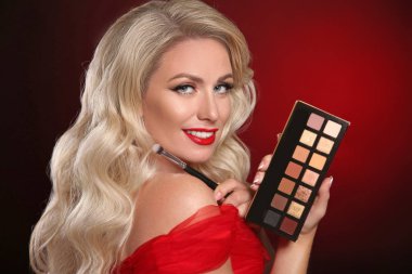 Güzellik makyajı. Kırmızı dudaklar, kozmetik göz farı paleti. Uzun dalgalı saç stili koyu arka planda ruj tutan seksi sarışın kadının moda portresi. Güzel kadın yüzü. 