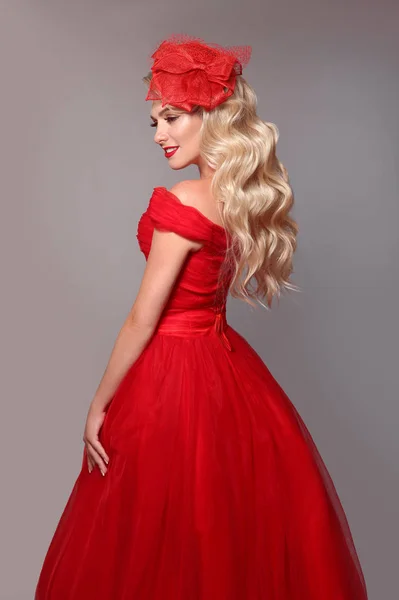 スタジオグレーの背景に隔離された赤いドレスとエレガントな帽子のブロンドの女性 長い波状の髪とメイクと美しいブロンドの女の子 花嫁のスタイル — ストック写真